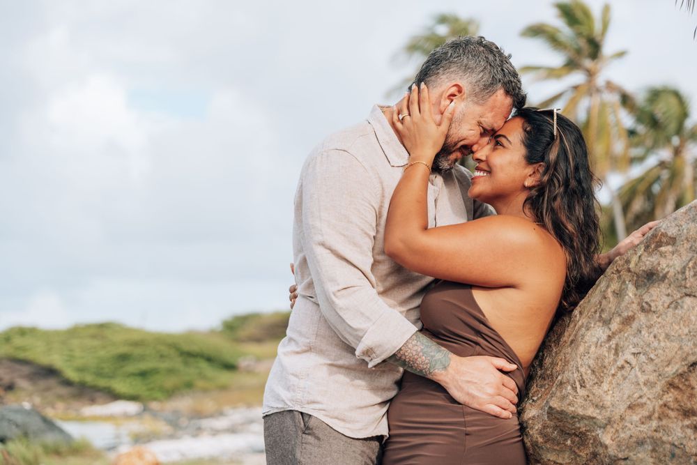 A romantic couple embraces near rocks and palm trees on a tropical beach during a cloudy day.