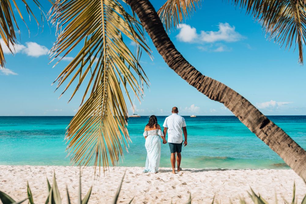 A couple stands on a pristine turquoise beach under leaning palm fronds in a tropical paradise.