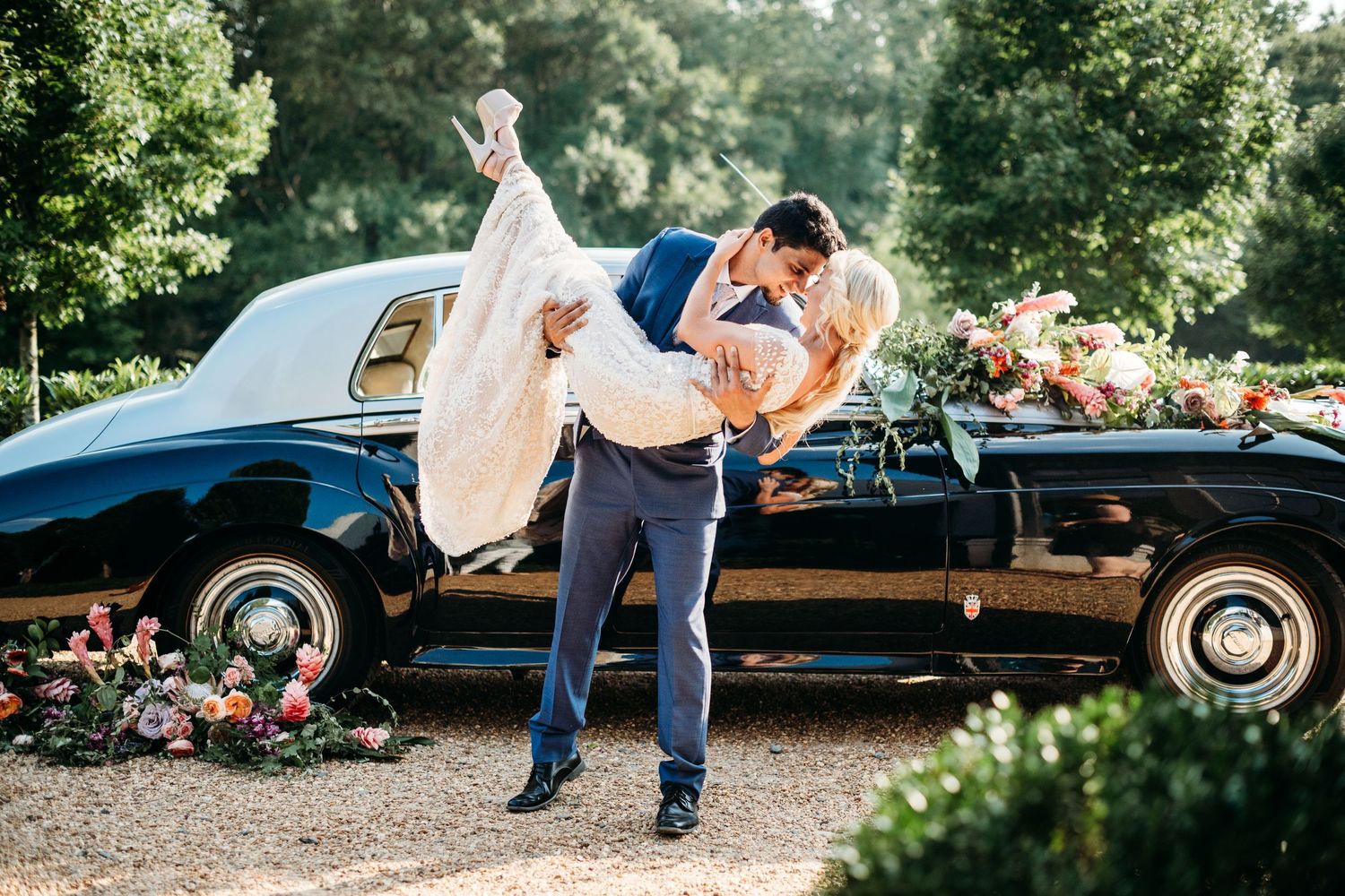 Groom lifts and dips his wife in front of an antique car with floral installations on the hood and by the wheel.