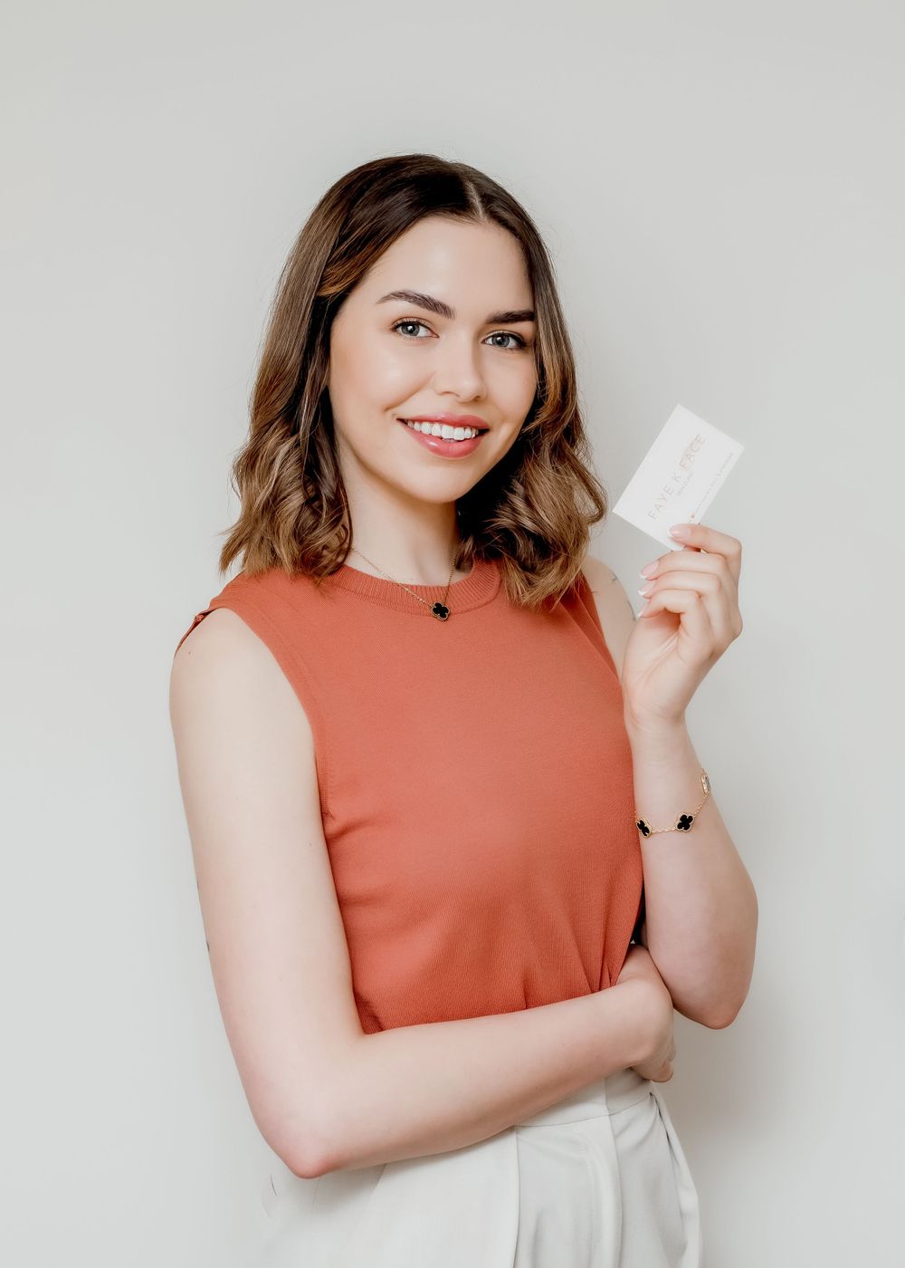 Model in coral sleeveless blouse and white trousers holding skincare product against light background.