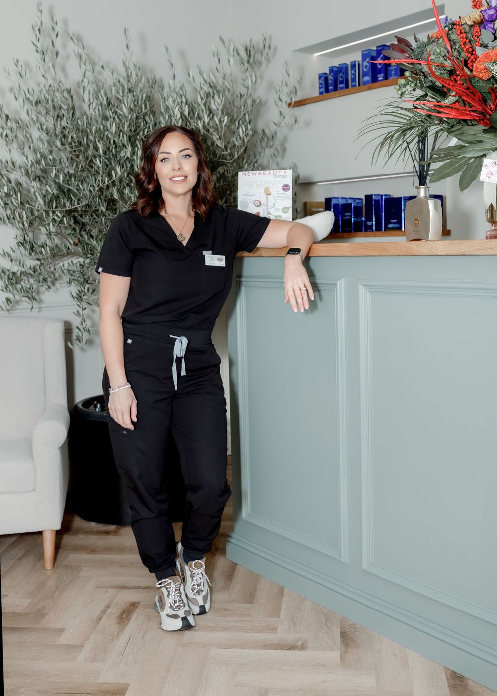 Healthcare professional in black uniform standing by counter with olive tree and floral arrangement.