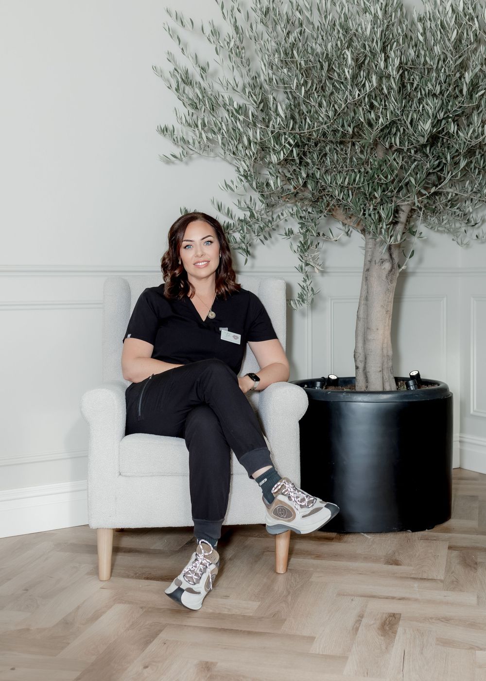 Healthcare worker relaxing in white armchair next to olive tree in modern office setting.