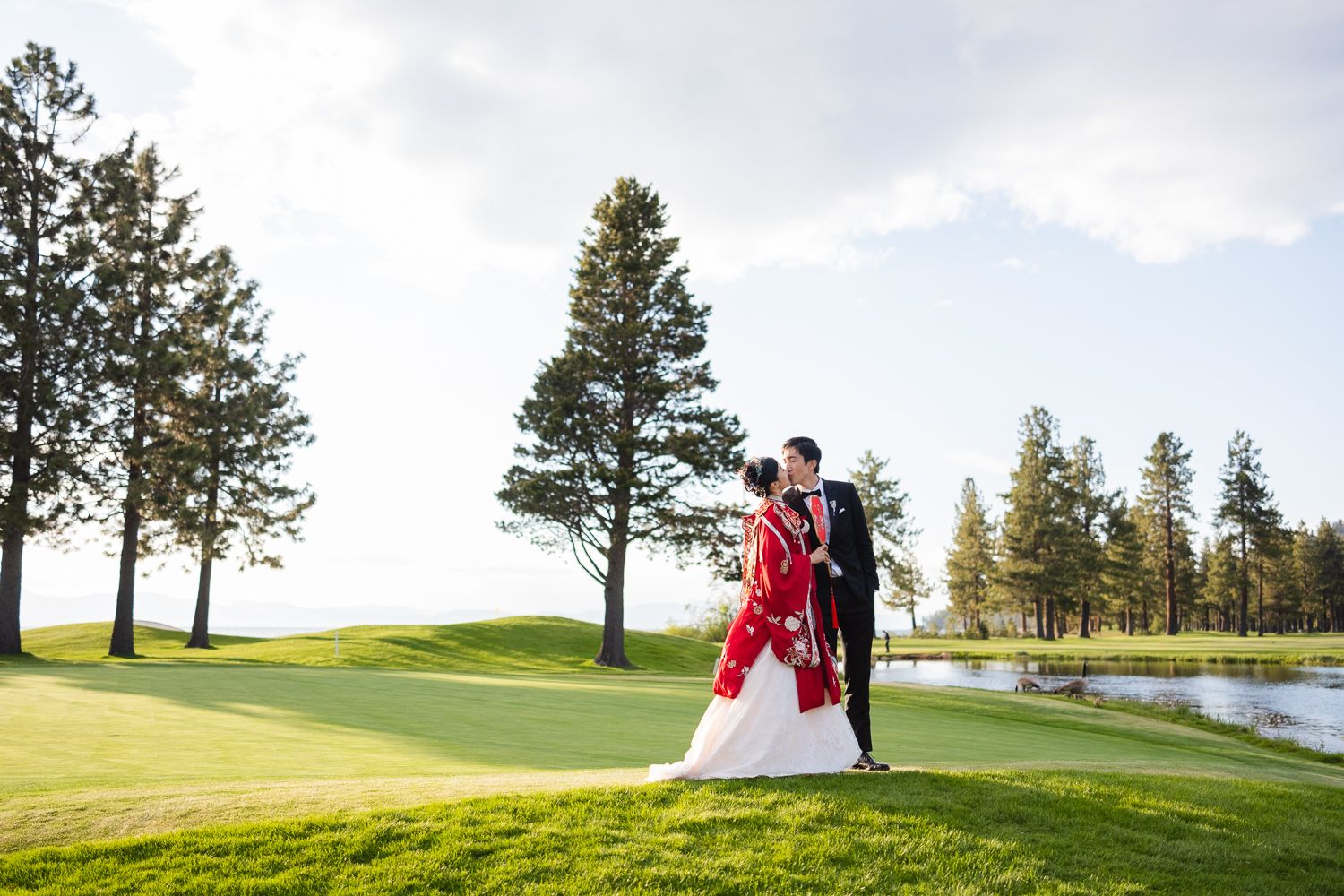 A couple in wedding attire embraces on a beautiful Edgewood golf course with tall pine trees and a pond in the background.