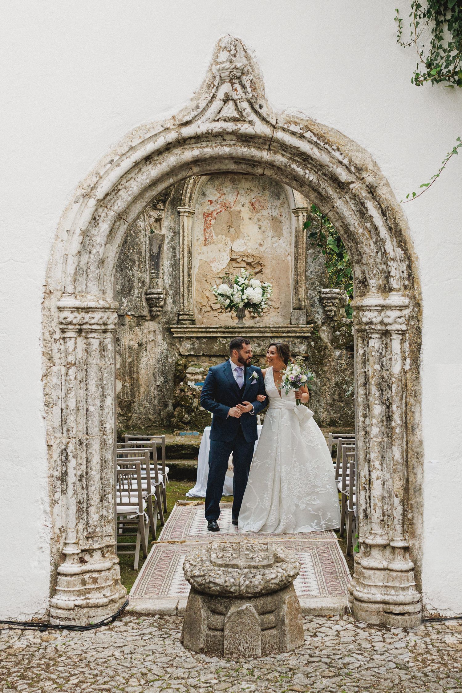 A wedding couple emerges through an ornate stone archway at a historic venue with a beautiful weathered architectural detail.