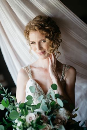 Portrait of a bride with curled hair holding a green and white bouquet against white fabric.