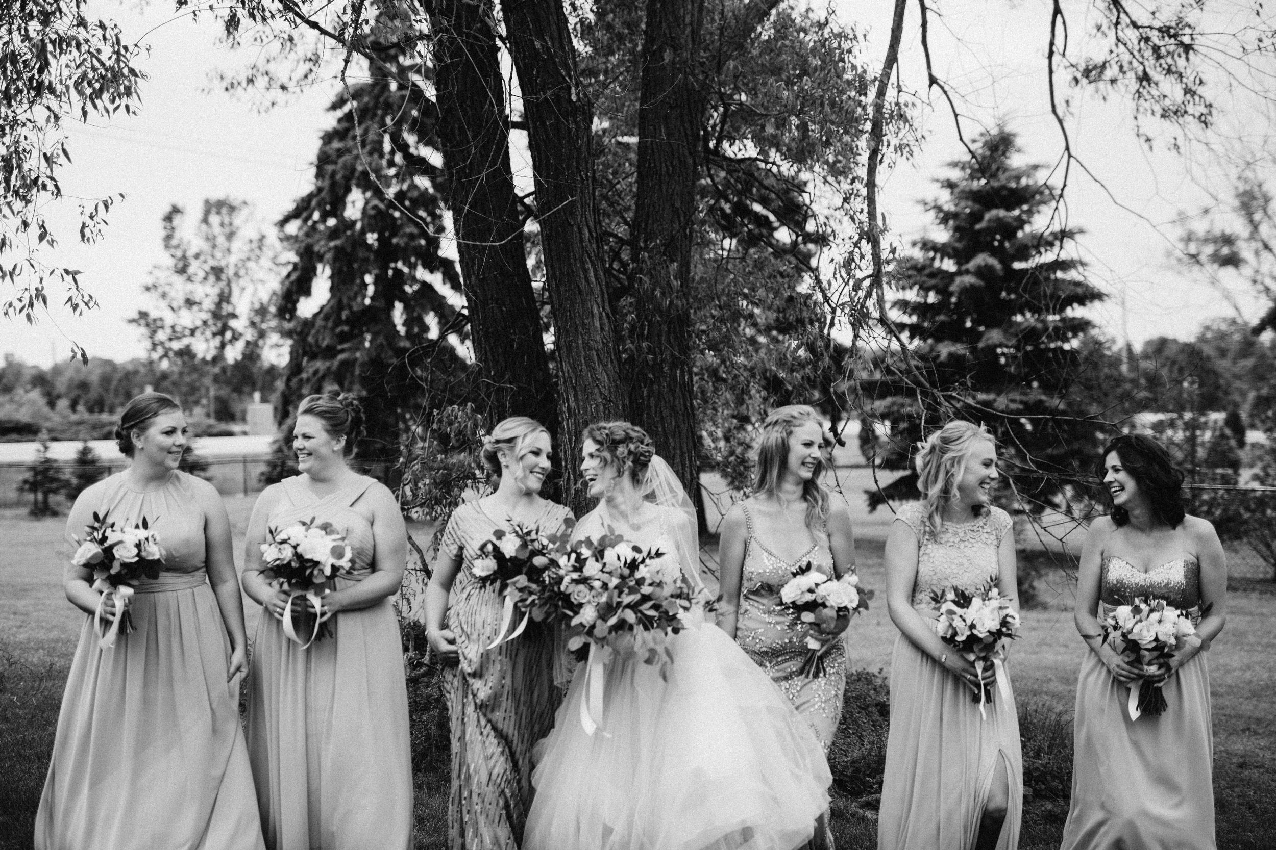 Wedding party standing under tall trees wearing light colored dresses and holding bouquets.
