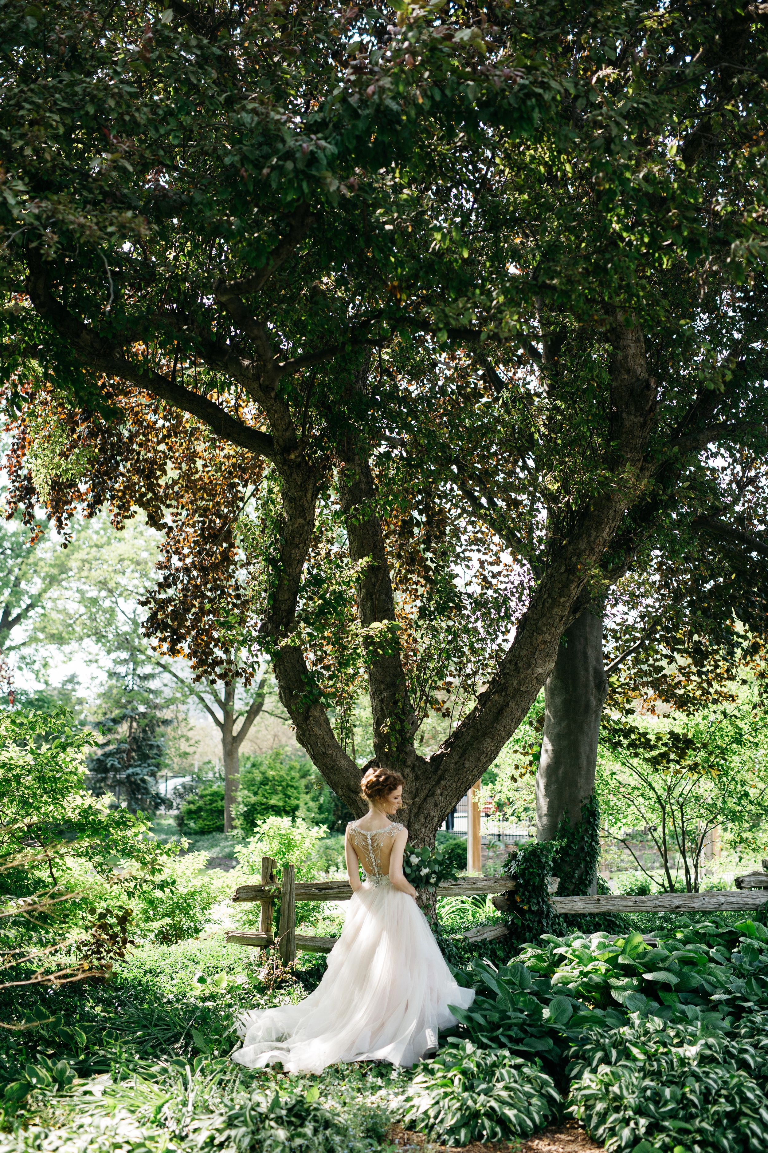 Bride in flowing white gown poses under a large tree in a garden setting.