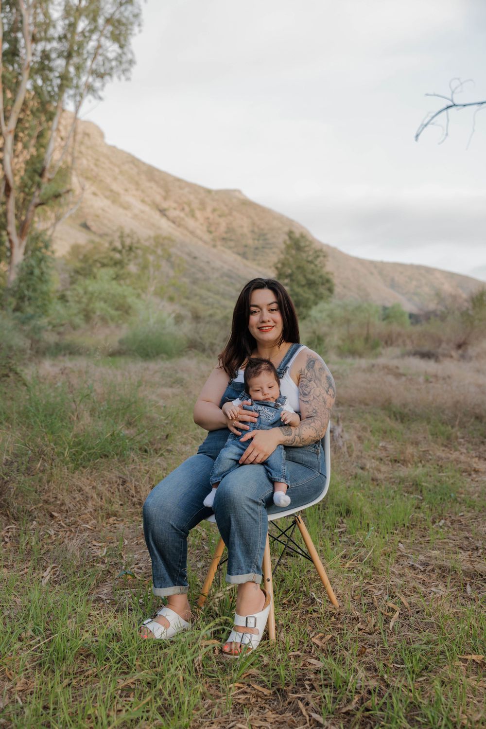 A seated figure with an infant sits on a modern chair in a scenic mountain landscape with trees on an overcast day.