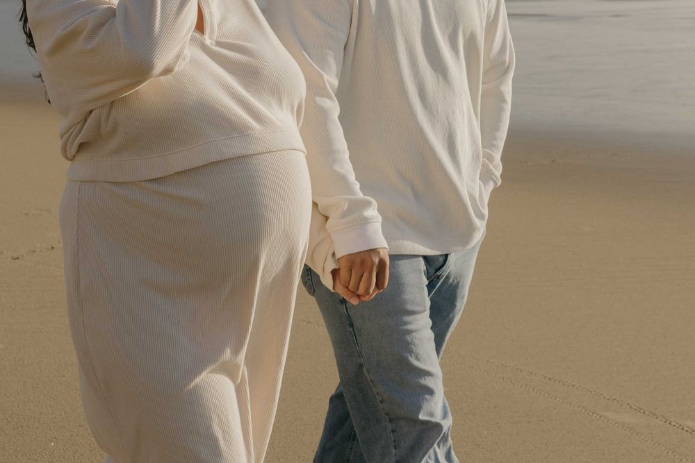 Two people walk on a sandy beach with one wearing a flowy cream dress and the other in light blue jeans.
