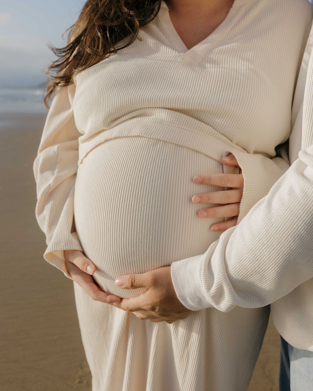 Pregnant woman in white sweater holding her belly tenderly while standing on beach at sunset.