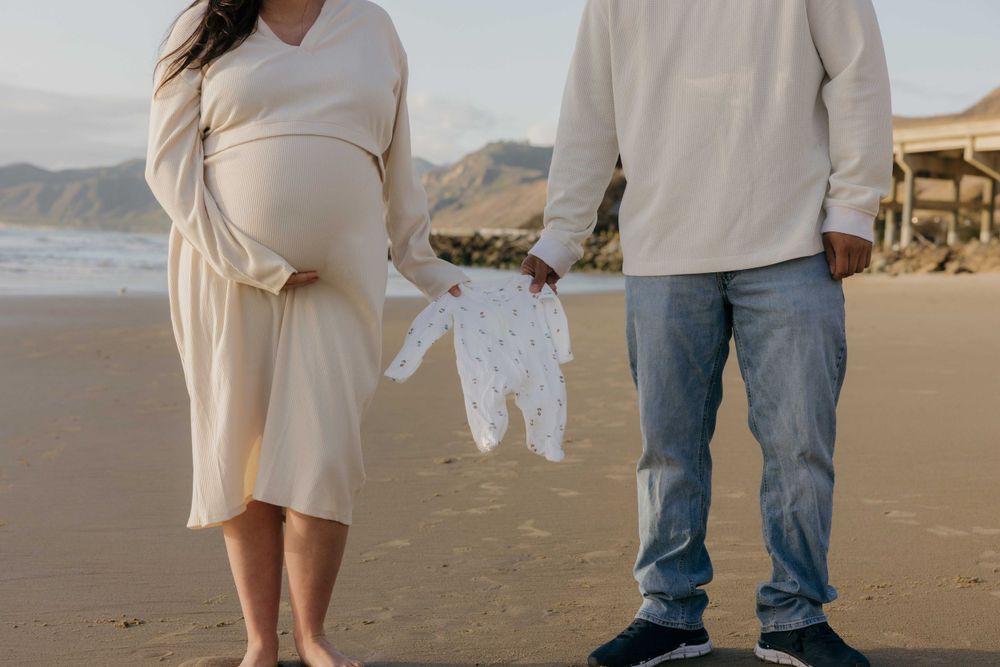 Expecting couple holds tiny white baby onesie while standing together on sandy beach at sunset.