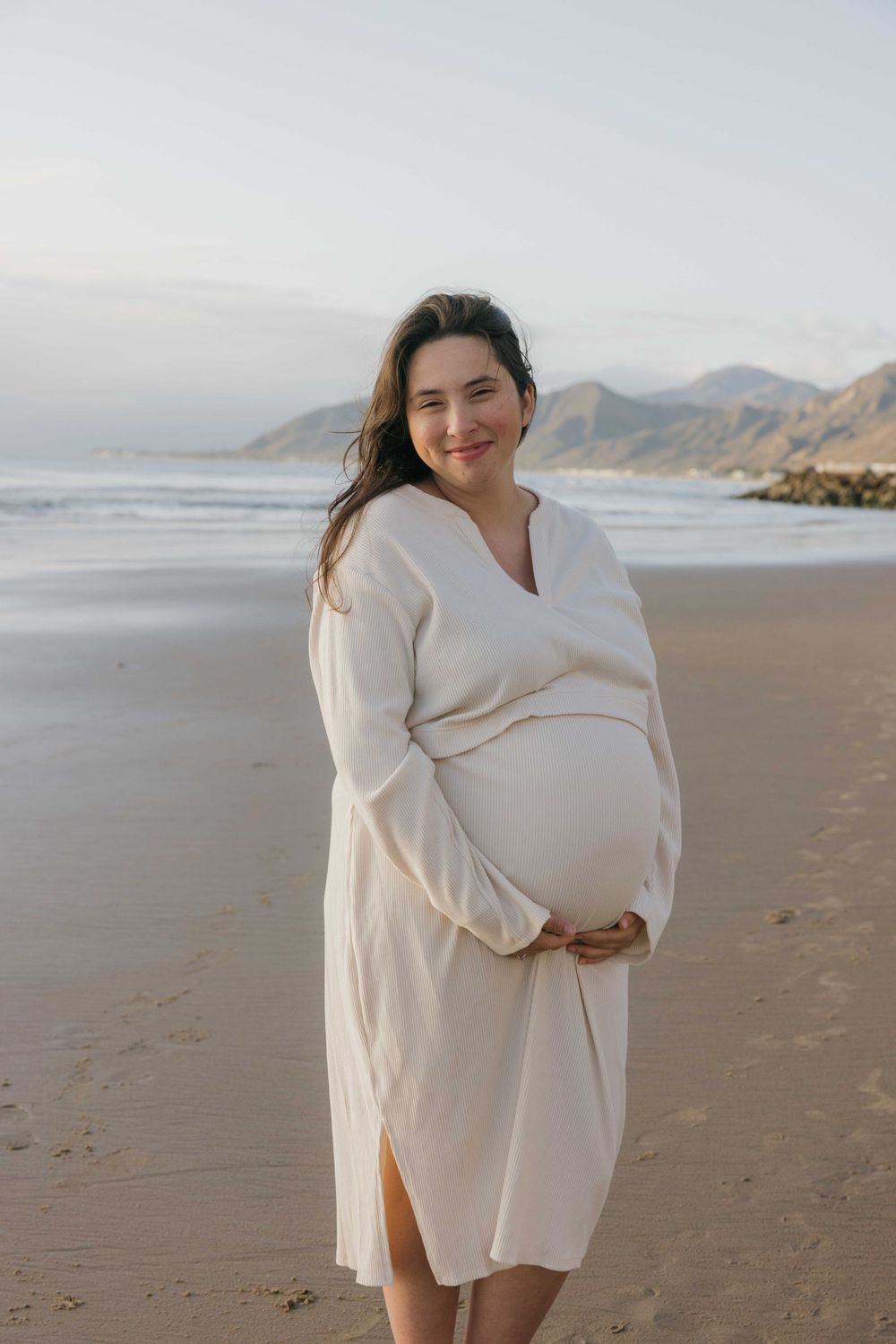 Expectant woman in white dress stands on peaceful beach at sunset with mountains in background.