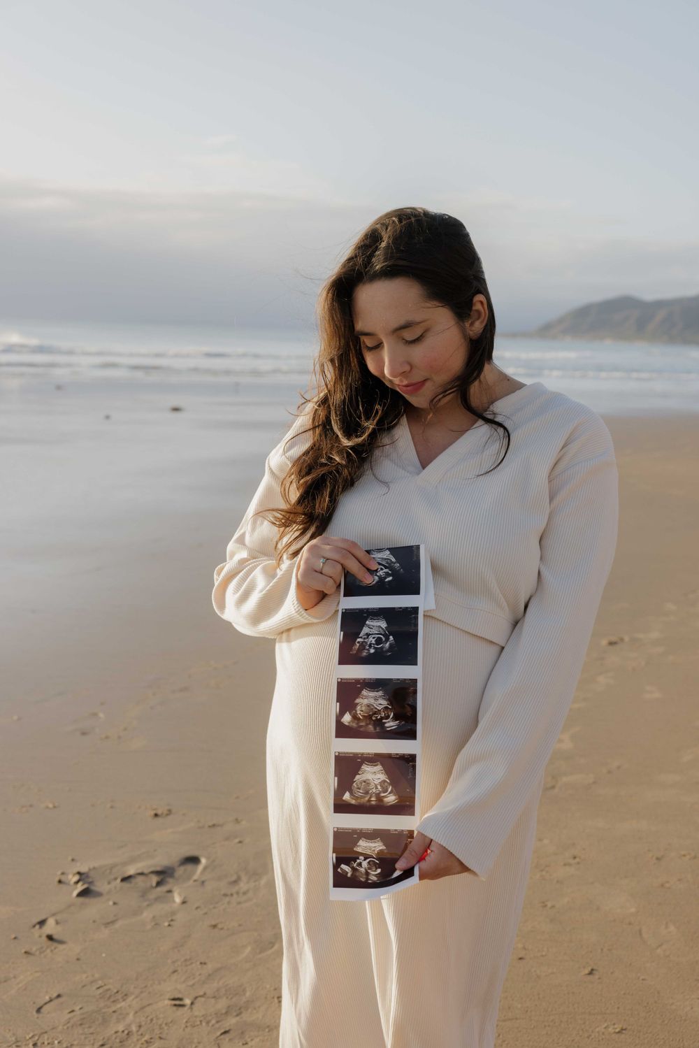 Someone wearing a cream-colored dress holds ultrasound photos while standing on a beach at sunset.