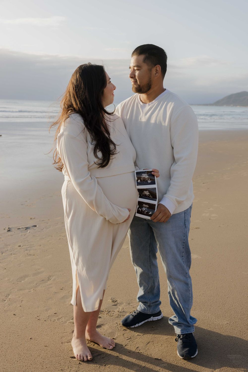 A couple in matching white sweaters stands on a beach holding ultrasound photos during a pregnancy announcement photo shoot.