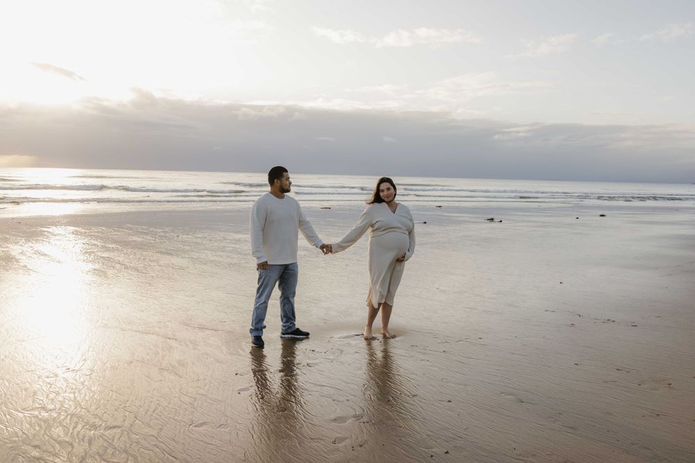 A couple holds hands while walking on a sunlit beach at sunset with waves and clouds in the background.