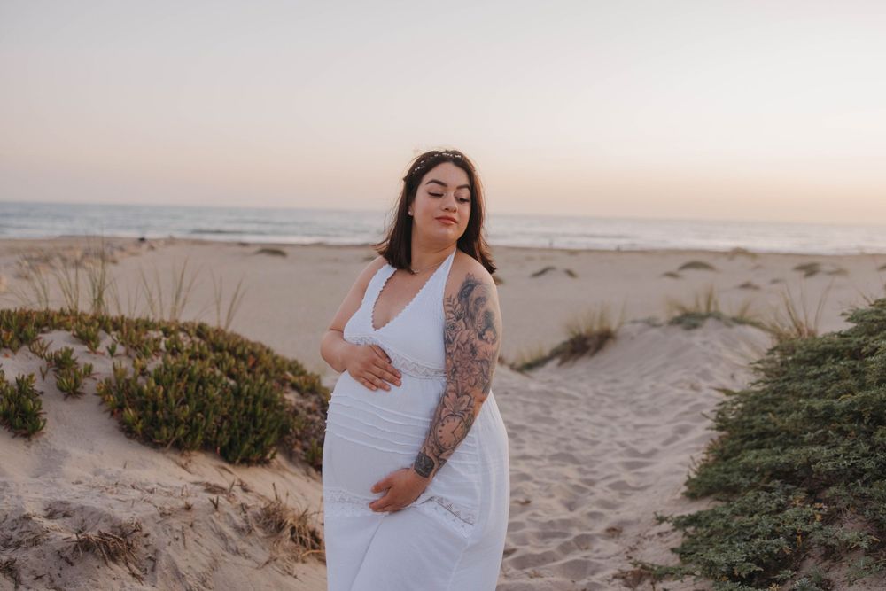 Expecting parent in flowing white dress poses on sandy beach path at sunset with scenic coastal background.