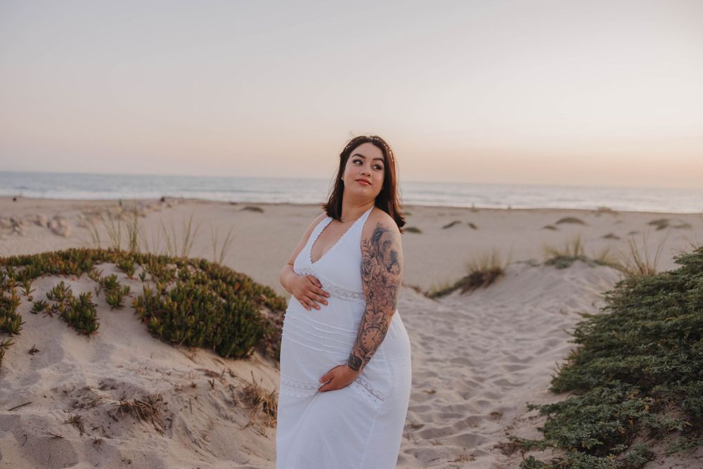 A pregnant woman in a white dress stands on a sandy beach path at sunset while holding her baby bump.