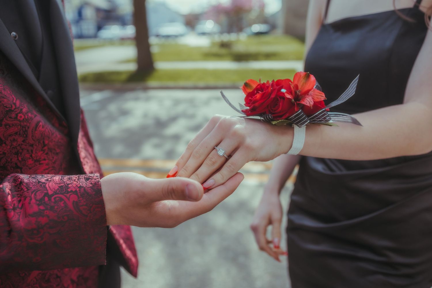 Close-up of hands exchanging red carnation corsage in outdoor setting.