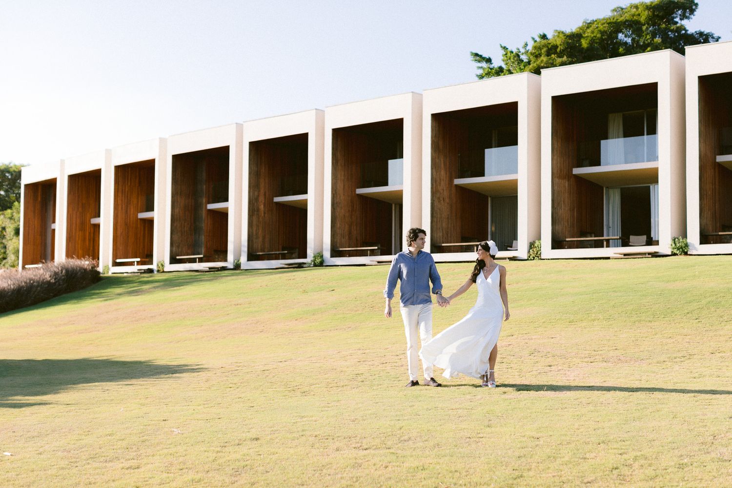 Two people holding hands walk across a grassy lawn in front of a modern building with geometric archways.