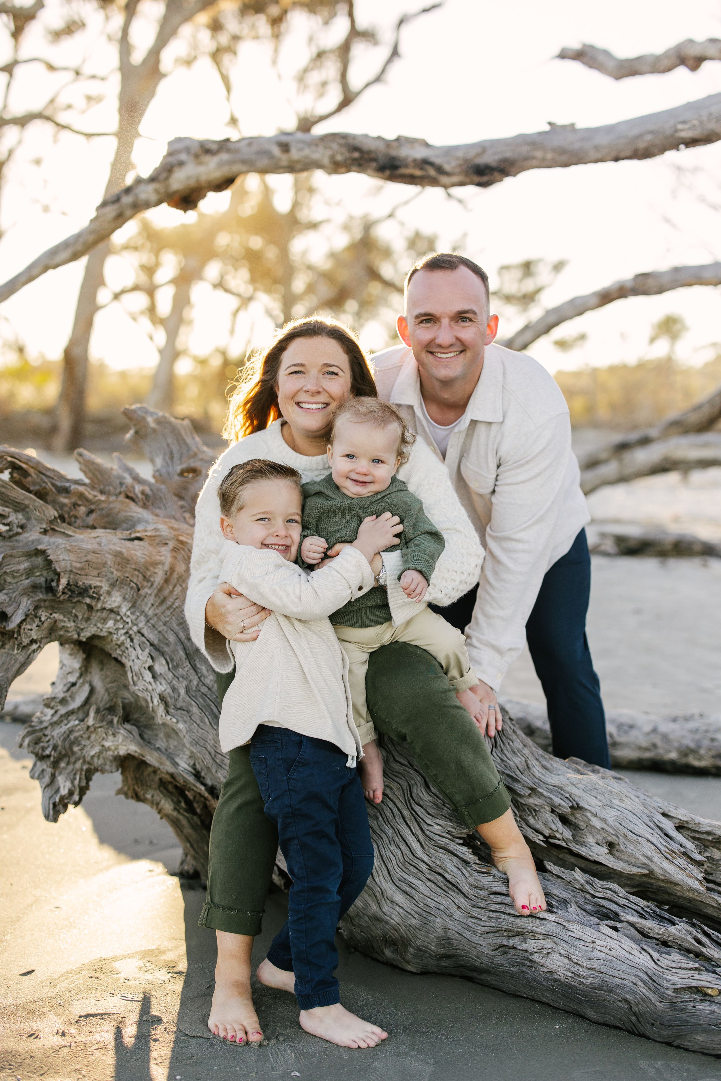 Family of four playing and laughing while sitting on driftwood at the beach during sunset.