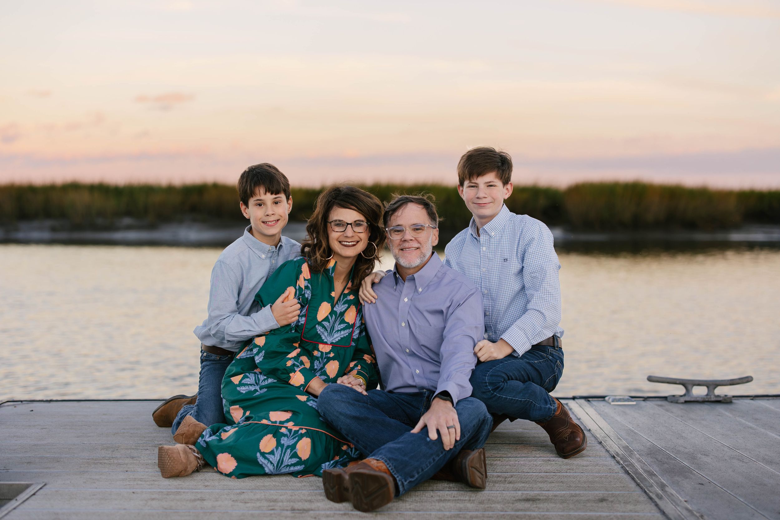 Family wearing coordinated blue and green outfits sits together on a dock at sunset near a lake.