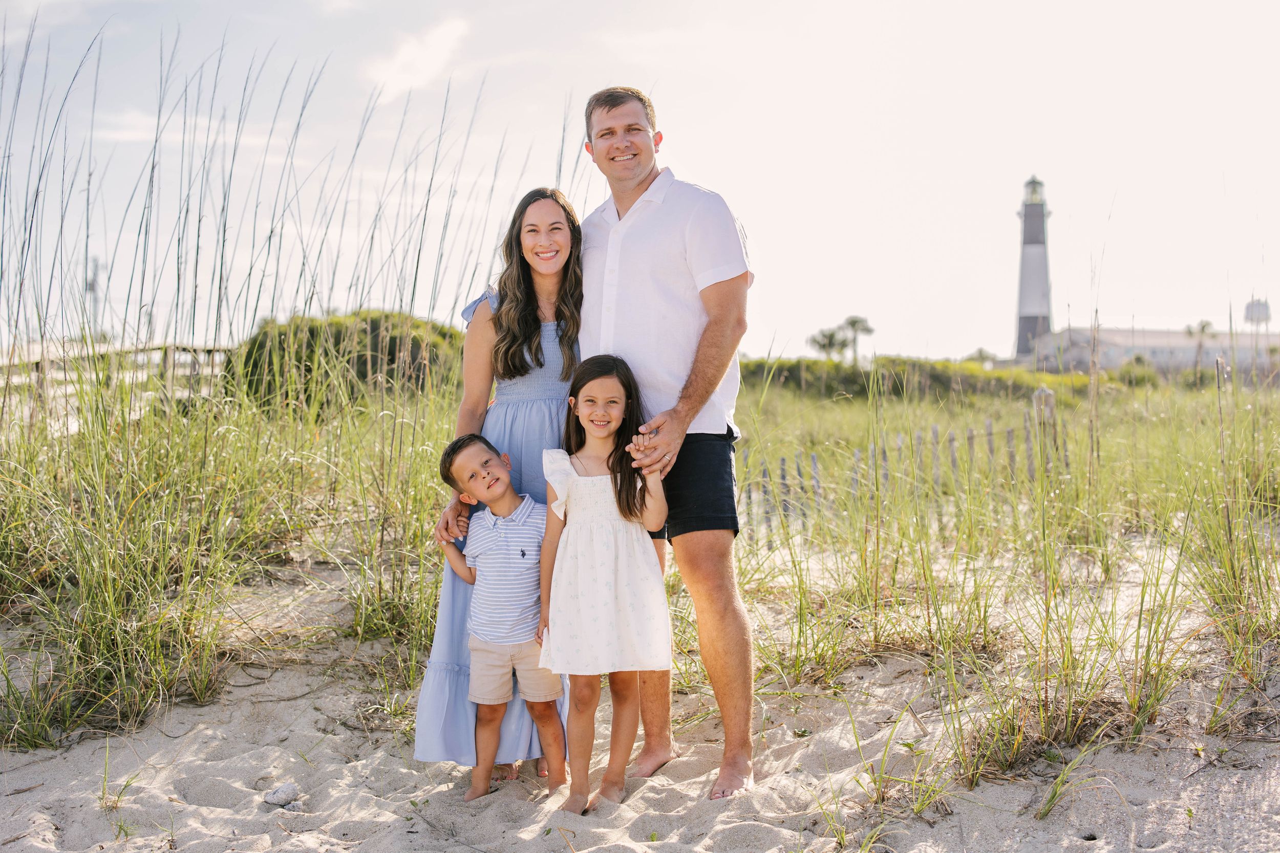 A group of people walk together along a sandy beach path through sea oats with a lighthouse visible in the background.