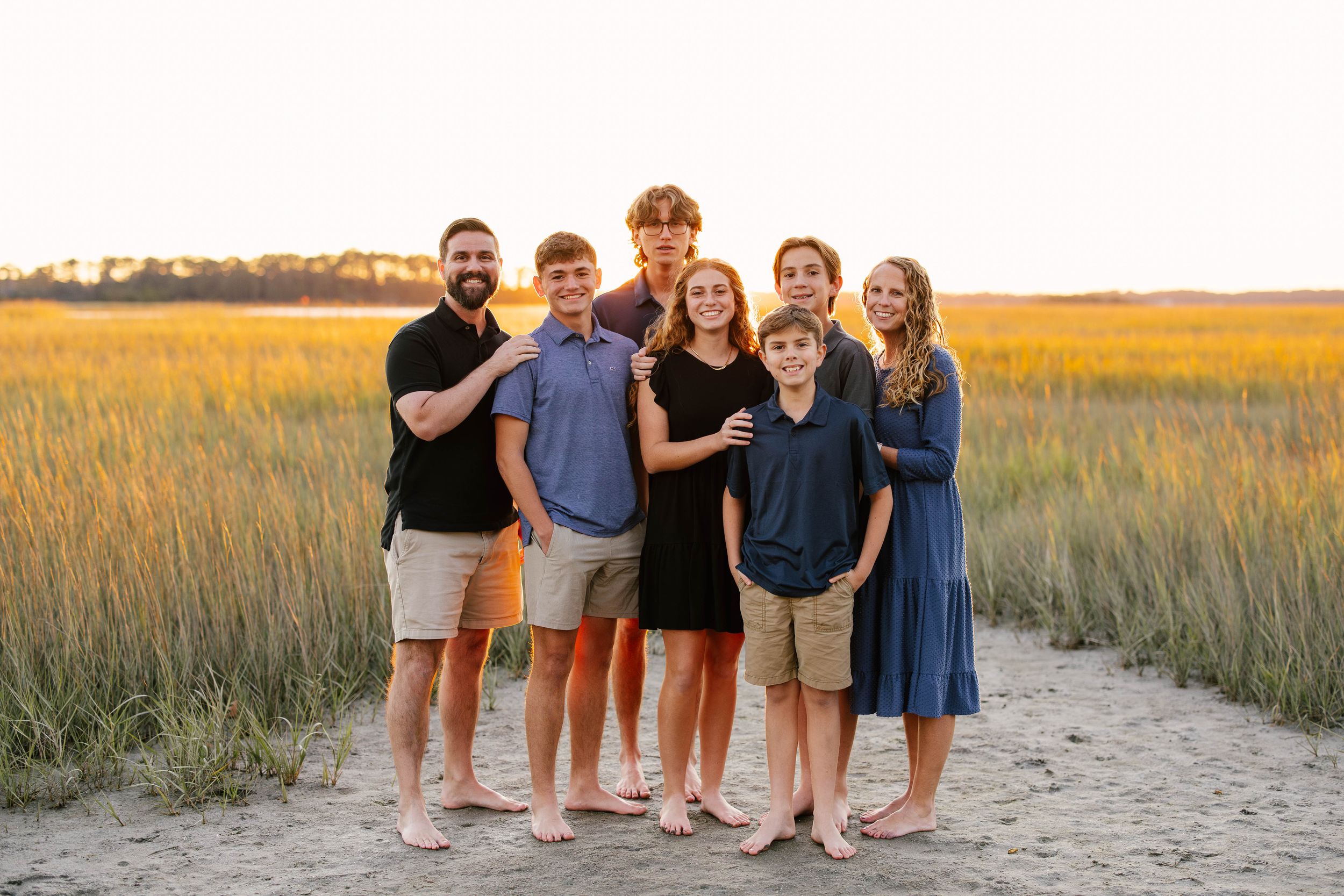 A group of people stand together on a sandy path through golden marsh grass at sunset.