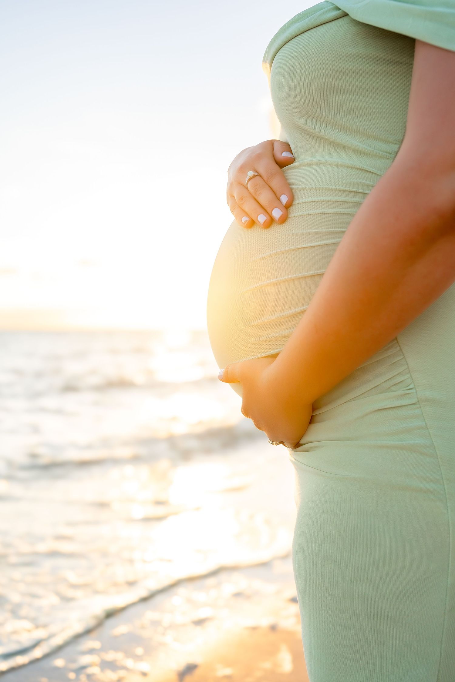 Close-up of pregnant woman in sage green dress cradling baby bump at golden hour beach.