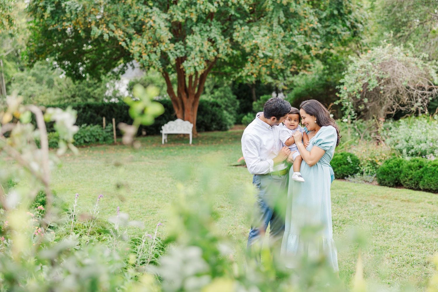 A couple stands together holding their baby in a lush garden setting with green trees and bushes in the background at Historic Tuckahoe