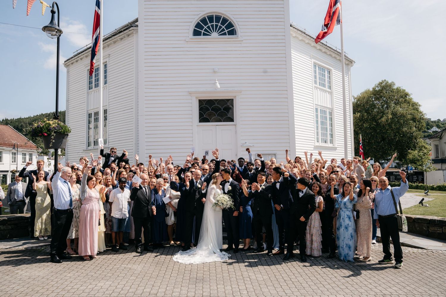 Large wedding group photo with guests cheering outside a white church on a sunny day.