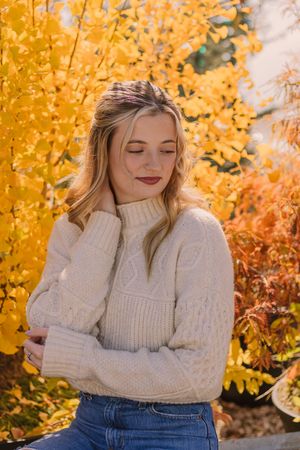 Medium close up of blonde girl wearing a white sweater in front of yellow tree.