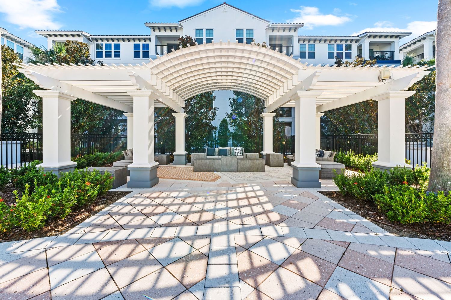 Elegant white stone pergola with arched walkway leads to a luxury residential building through manicured gardens.