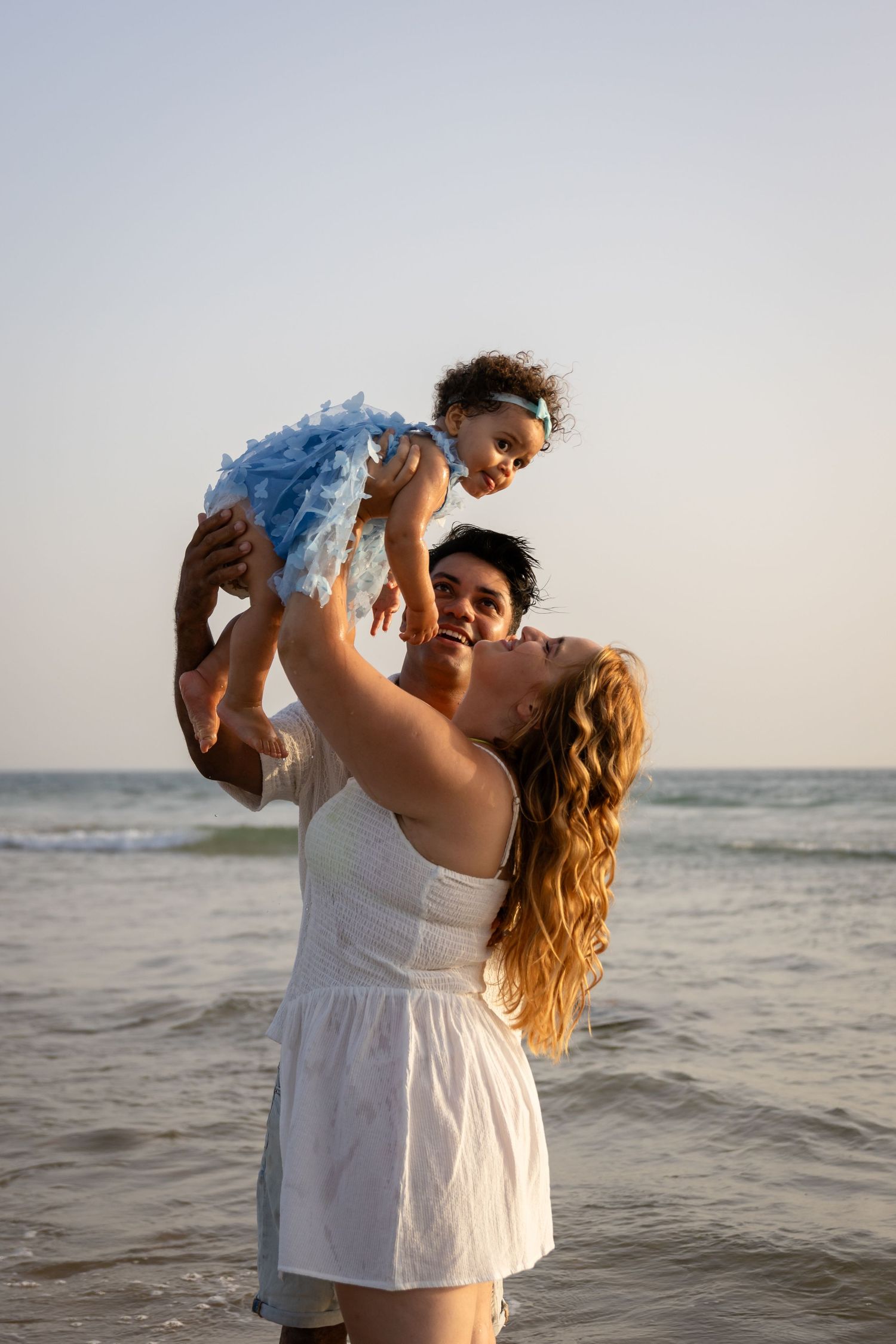 A couple in white clothing dances playfully together at the beach during sunset.
