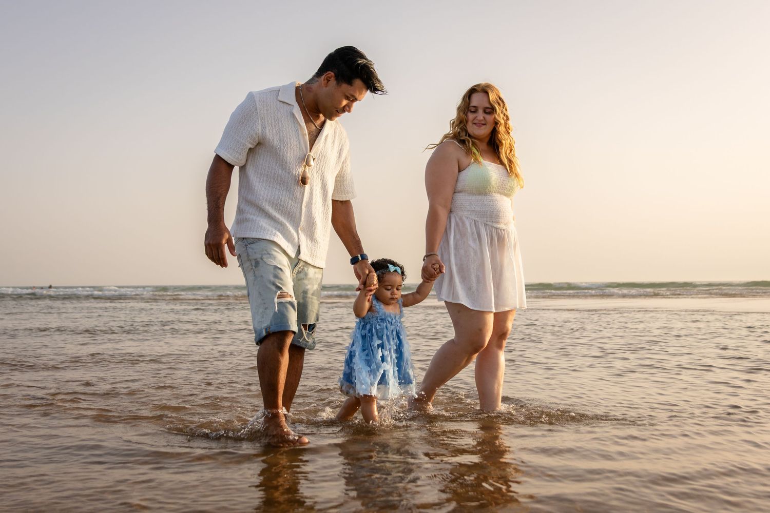 Two people wade through shallow ocean water at sunset wearing white and blue outfits.