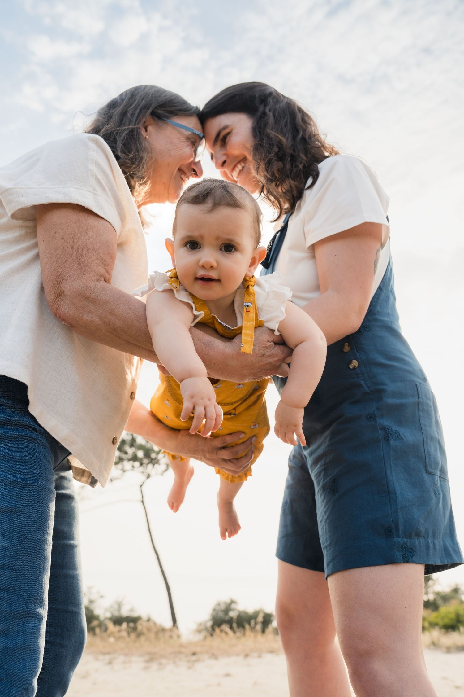 Two women holding a baby outdoors under a bright sky, sharing a tender forehead-to-forehead moment.
