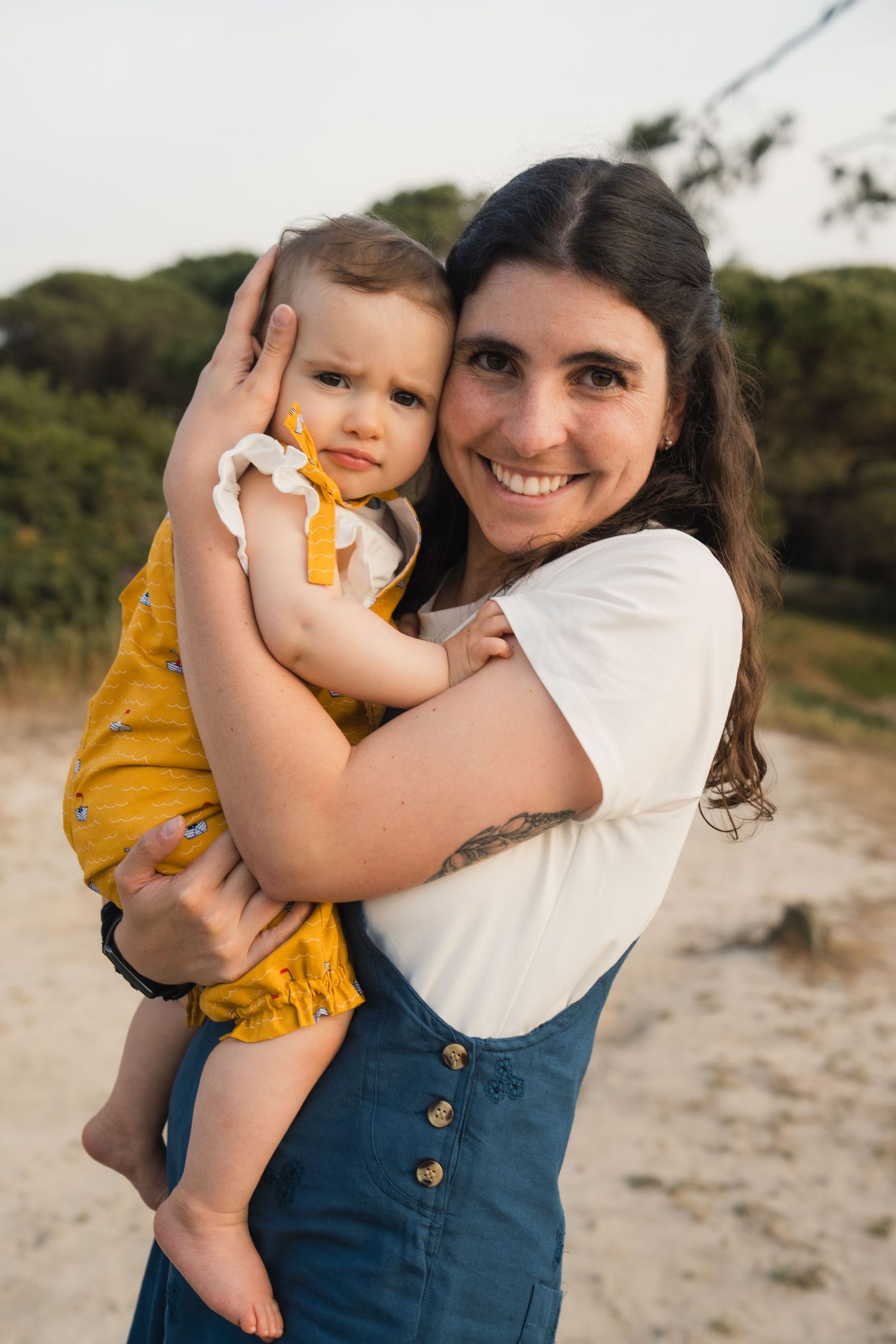 Smiling woman in white top and blue overalls holding a baby in yellow outfit outdoors.