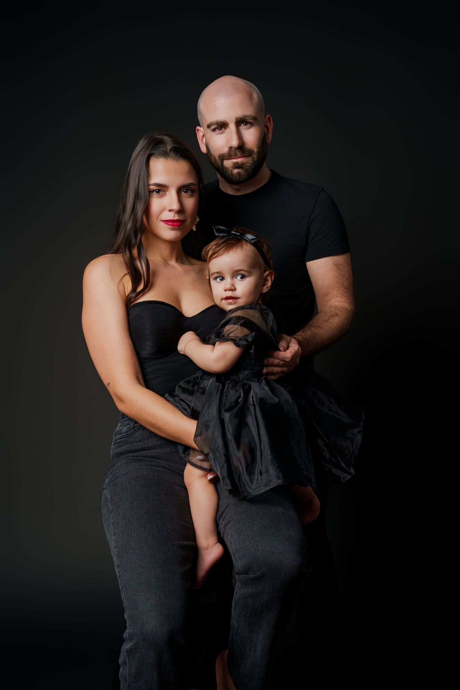 Family in black outfits poses together against dark dramatic background.