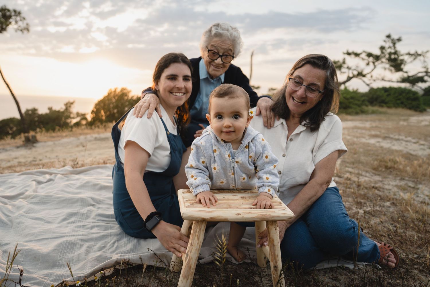 Three generations of women pose outdoors at sunset with a baby at a rustic wooden stool in a golden field.