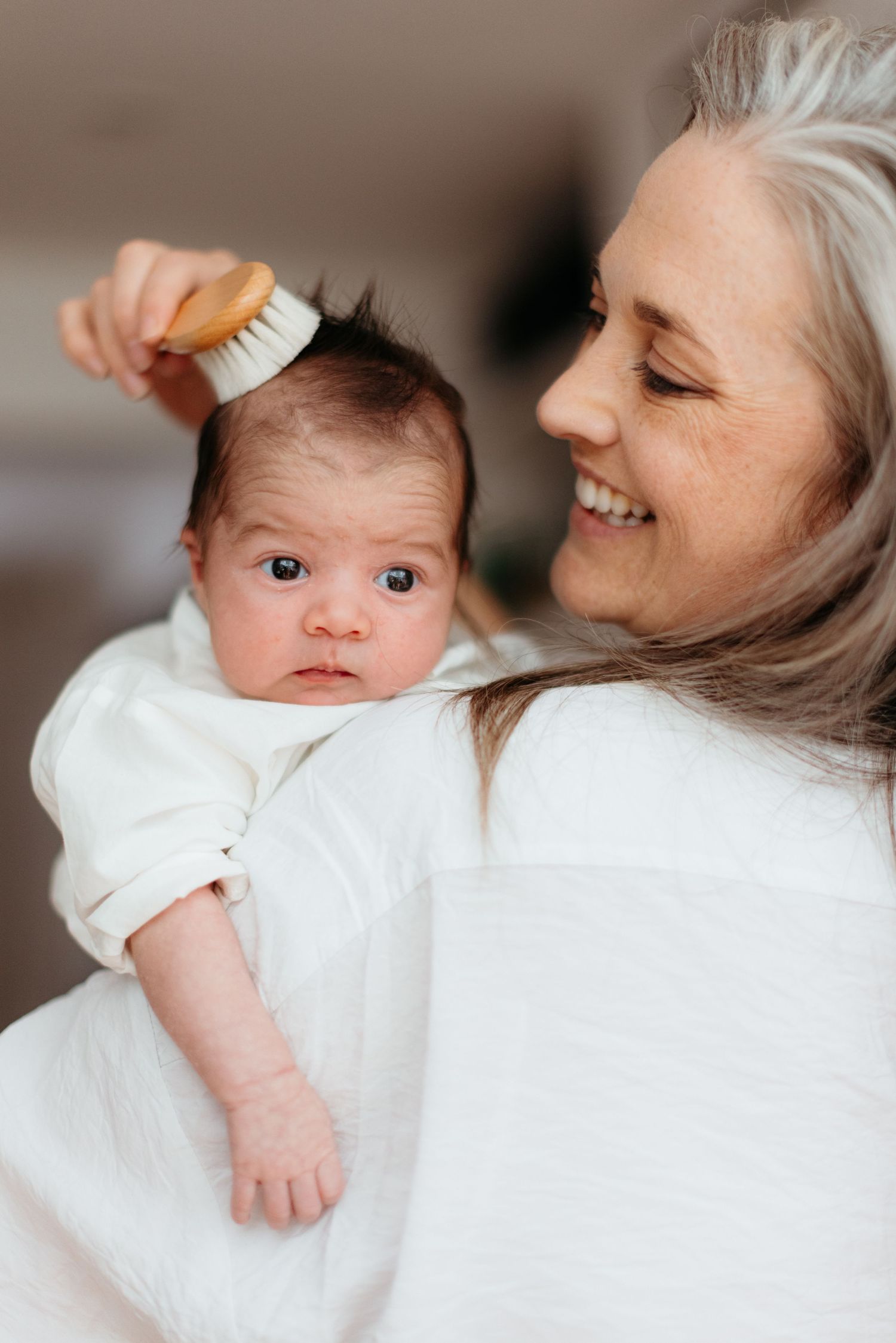 Parent gently brushing baby's dark hair while both wearing white clothing in warm intimate moment.