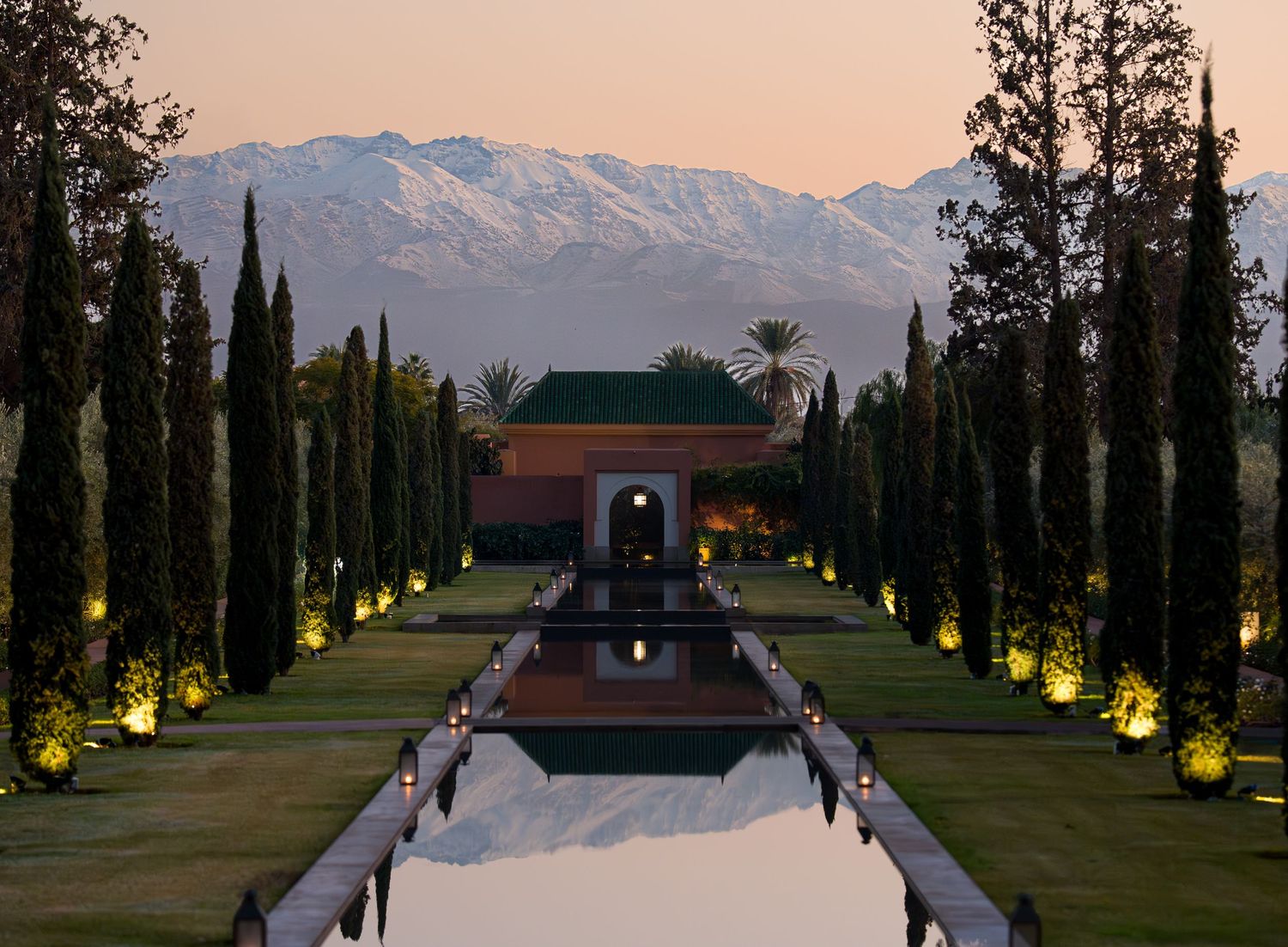 A scenic mountain resort venue with cypress trees lining a reflective pool at sunset against snowy peaks in the background.