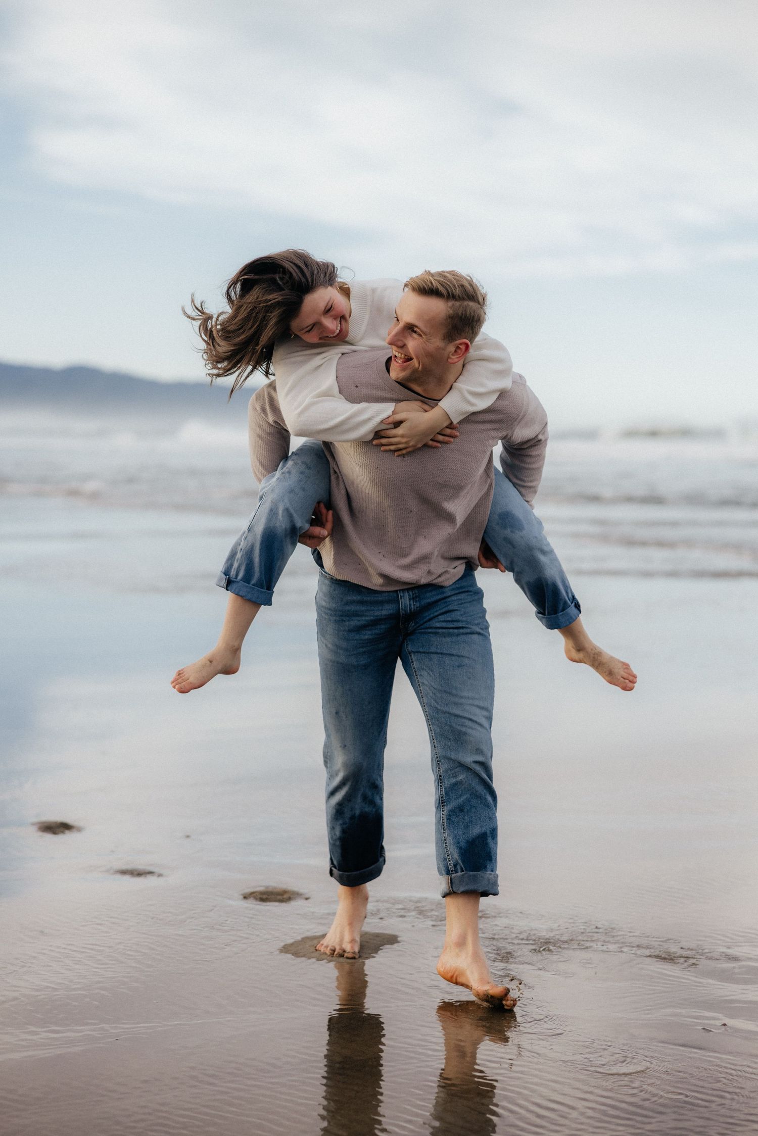 Couple having fun playing piggyback on a misty beach with mountains in the background.