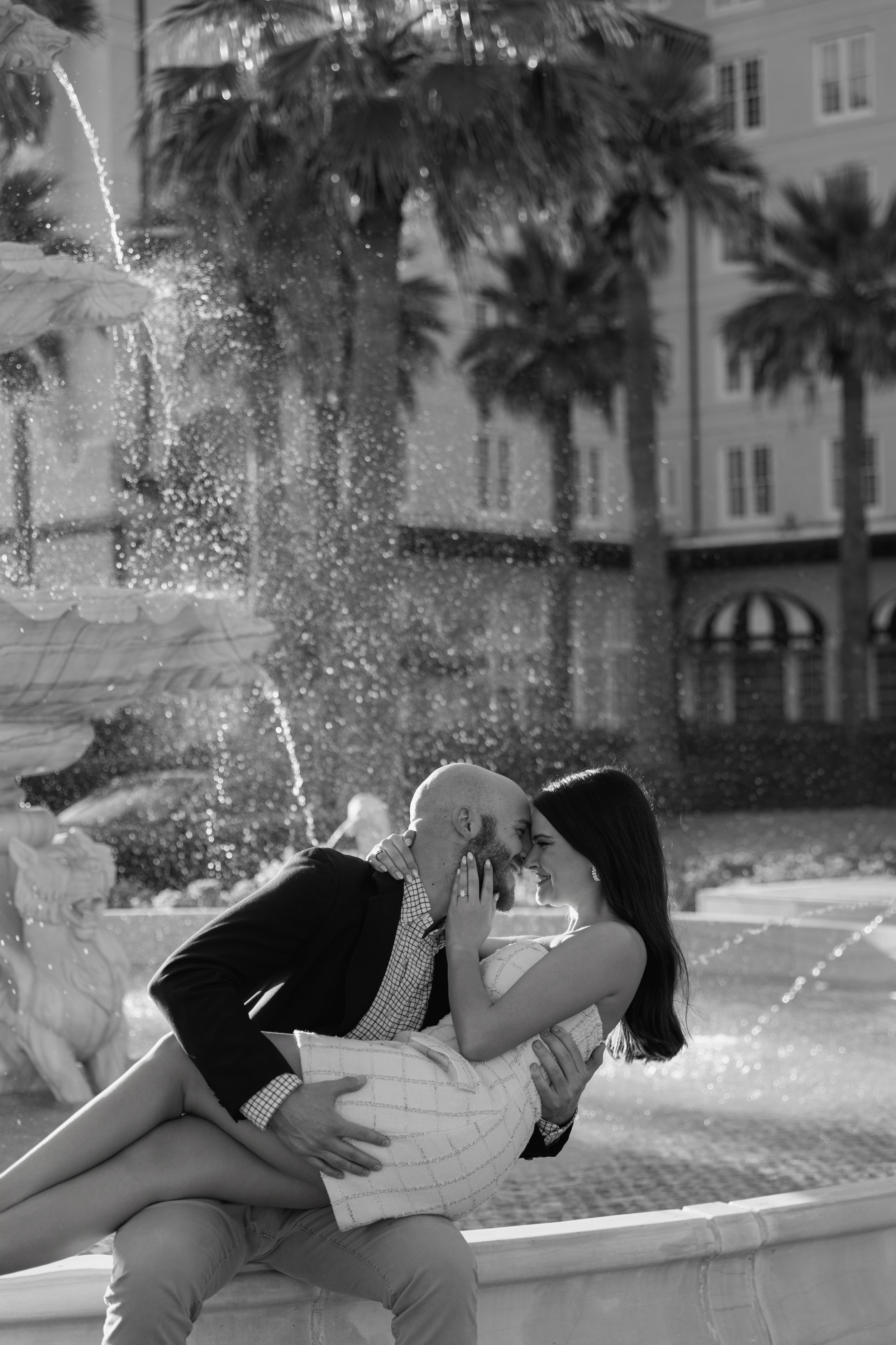 Black and white photo of couple kissing by ornate fountain with palm trees in background.
