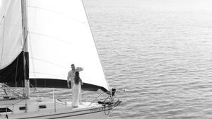 Black and white photo of couple standing on sailboat bow on calm open water.