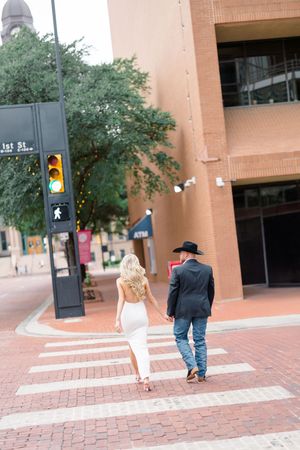 Couple holding hands crossing a brick crosswalk in downtown city street, woman in white dress and man in black cowboy hat.
