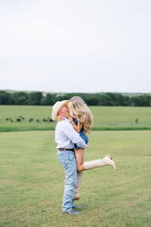 Man in cowboy hat lifting woman in denim outfit in a green pasture with cattle visible in the background.