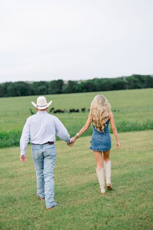 Couple walking hand in hand through green pasture, man in white cowboy hat and woman in denim skirt with cowboy boots.