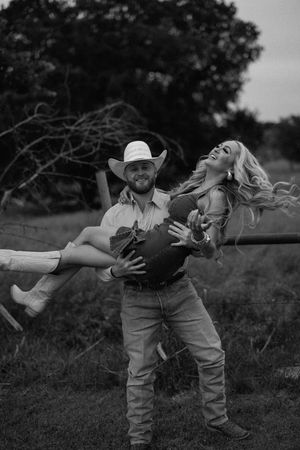 Black and white photo of smiling man in cowboy hat carrying laughing woman in his arms outdoors.