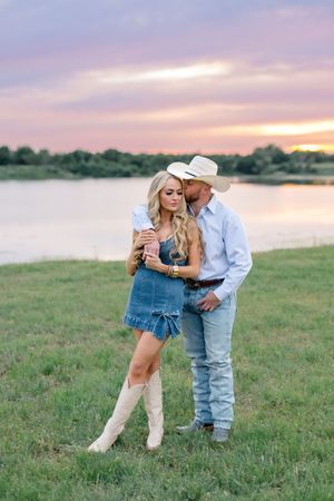 Couple embracing at sunset by a lake, woman in denim romper with white cowboy boots and man in cowboy hat.