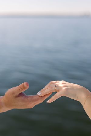 Close-up of two hands reaching toward each other, woman wearing a diamond engagement ring, with calm ocean water in background.