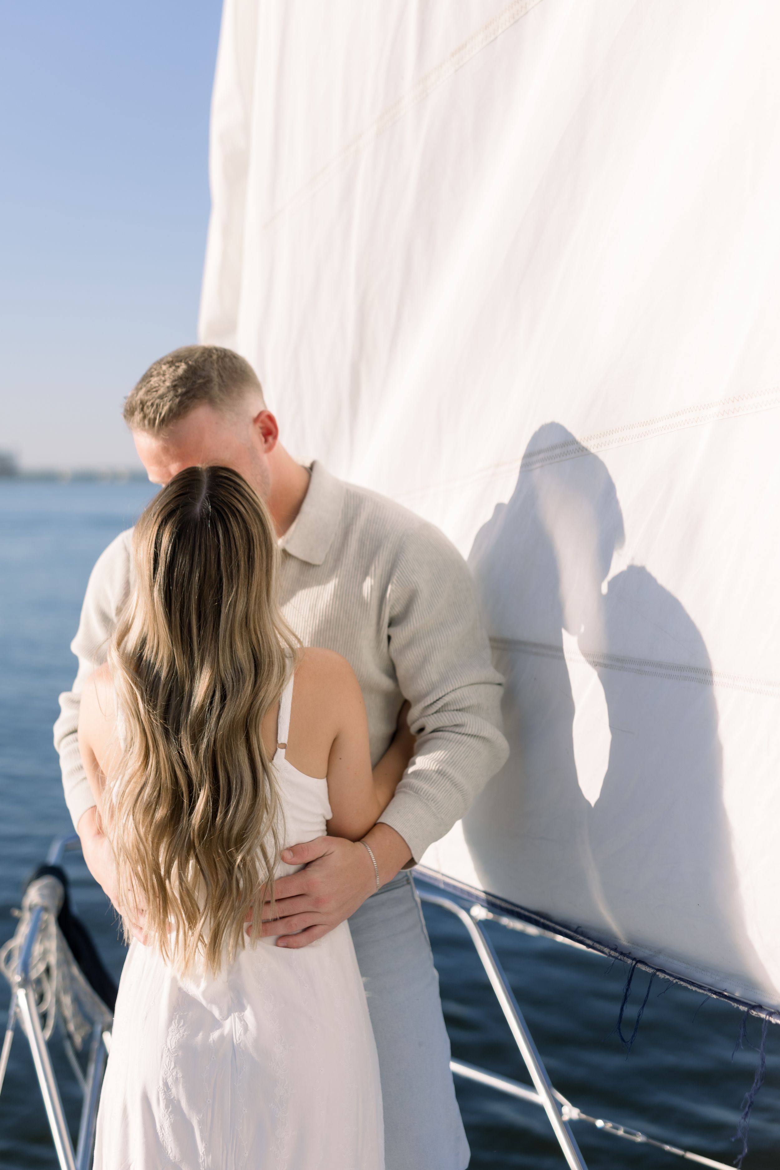 Couple sharing a romantic kiss on a sailboat deck, woman in white dress with long hair, man in beige sweater, blue sky background.